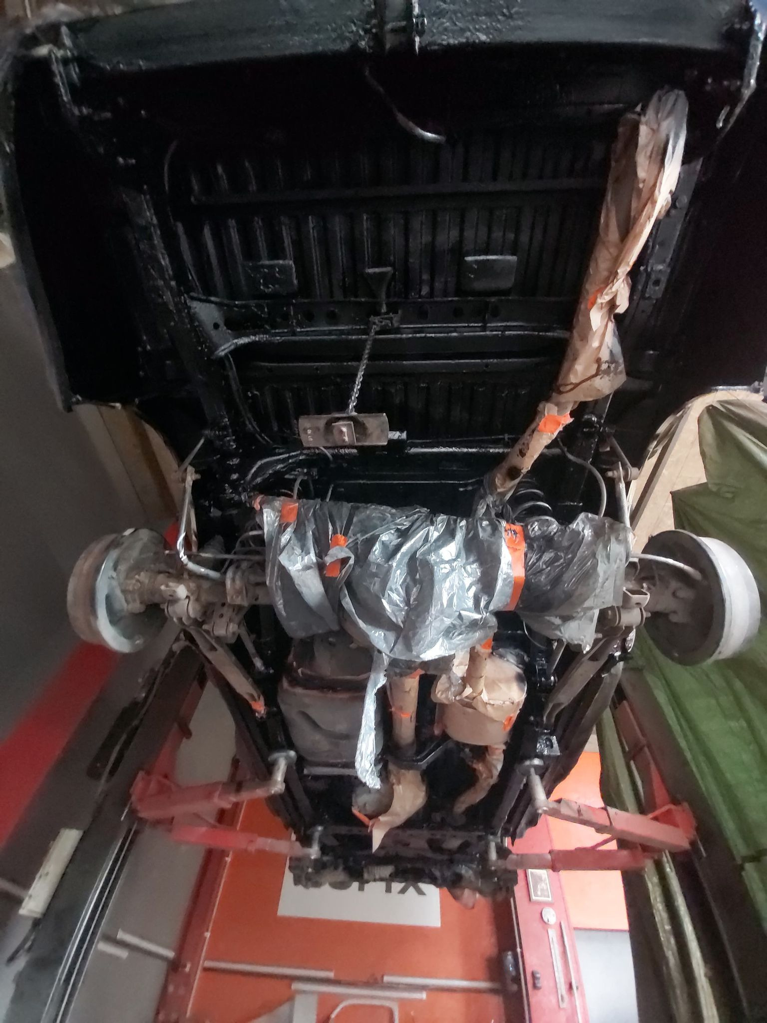 Podwozie samochodu na podnośniku, zabezpieczone folią i taśmą przed malowaniem, świeżo pokryte czarną farbą antykorozyjną.