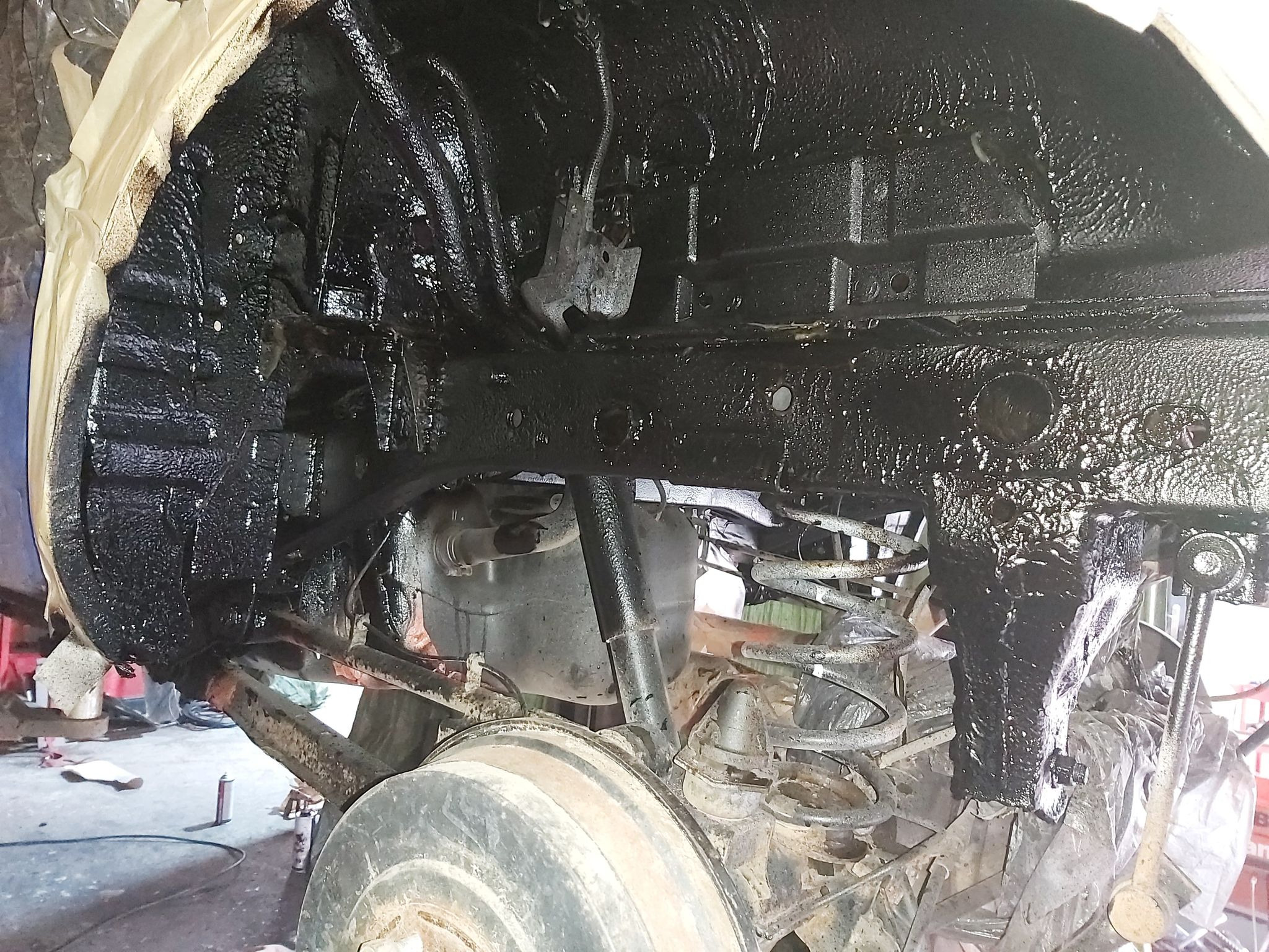 Podwozie samochodu po nałożeniu czarnej, ochronnej powłoki antykorozyjnej; widoczne elementy zawieszenia, sprężyny, amortyzatory i fragmenty karoserii zabezpieczone folią.