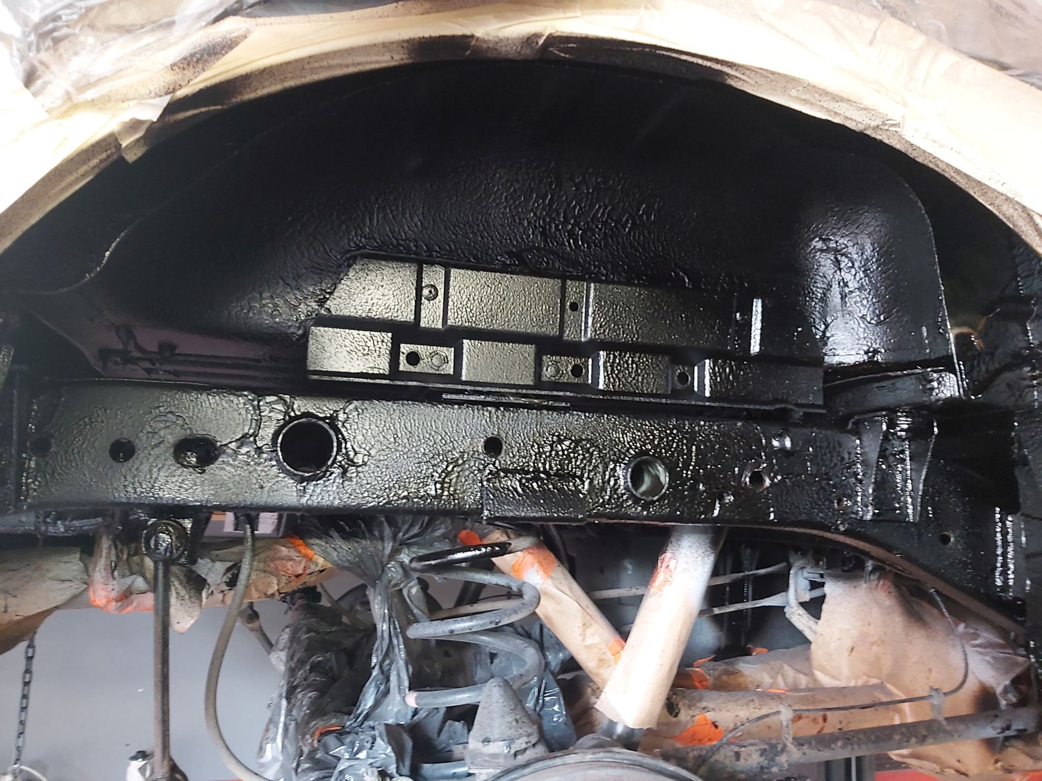 Podwozie samochodu po nałożeniu czarnej powłoki antykorozyjnej, widoczne elementy zawieszenia zabezpieczone folią i taśmą malarską.