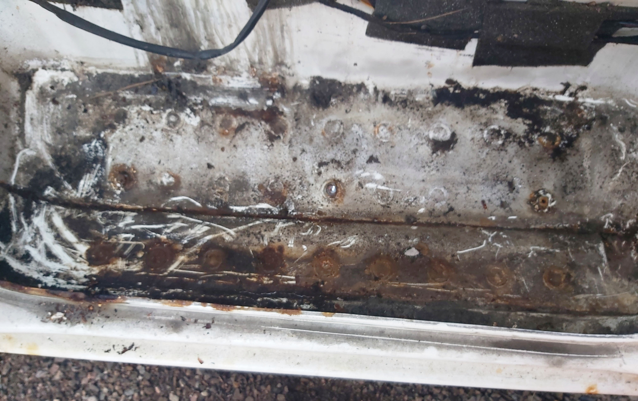 Wnętrze progu samochodu z widoczną korozją i śladami napraw, zarysowania na białej powierzchni, widoczne punkty spawania.