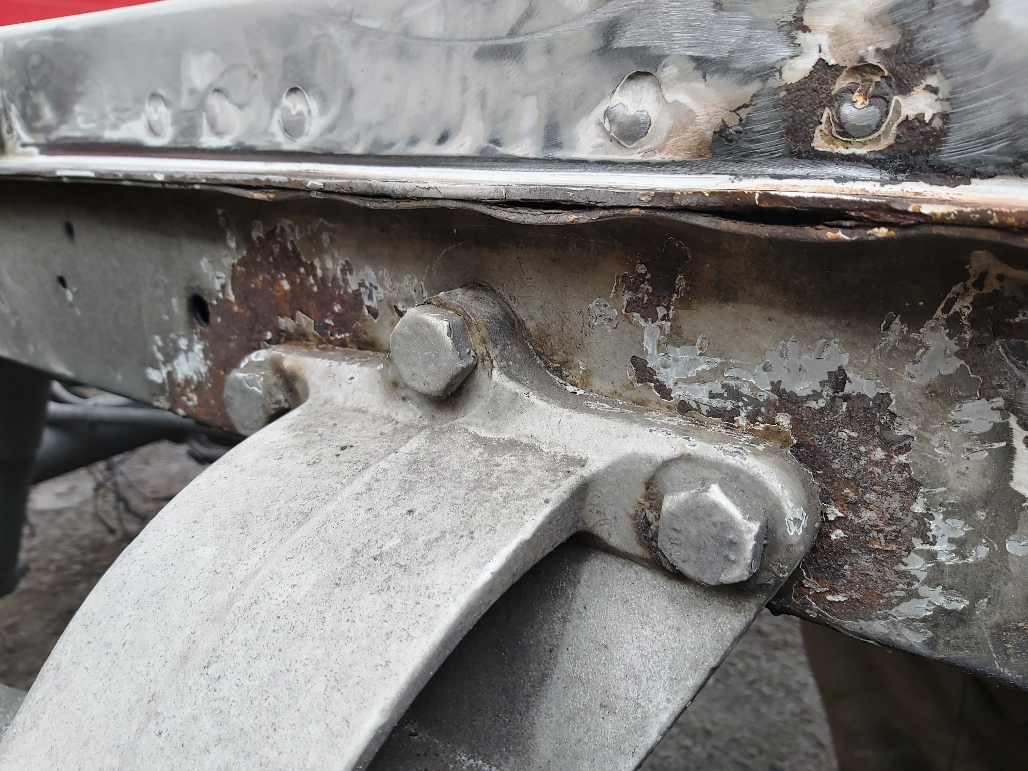 Fragment podwozia samochodu z widocznymi śladami korozji i napraw blacharskich, z elementami łączącymi na śruby.
