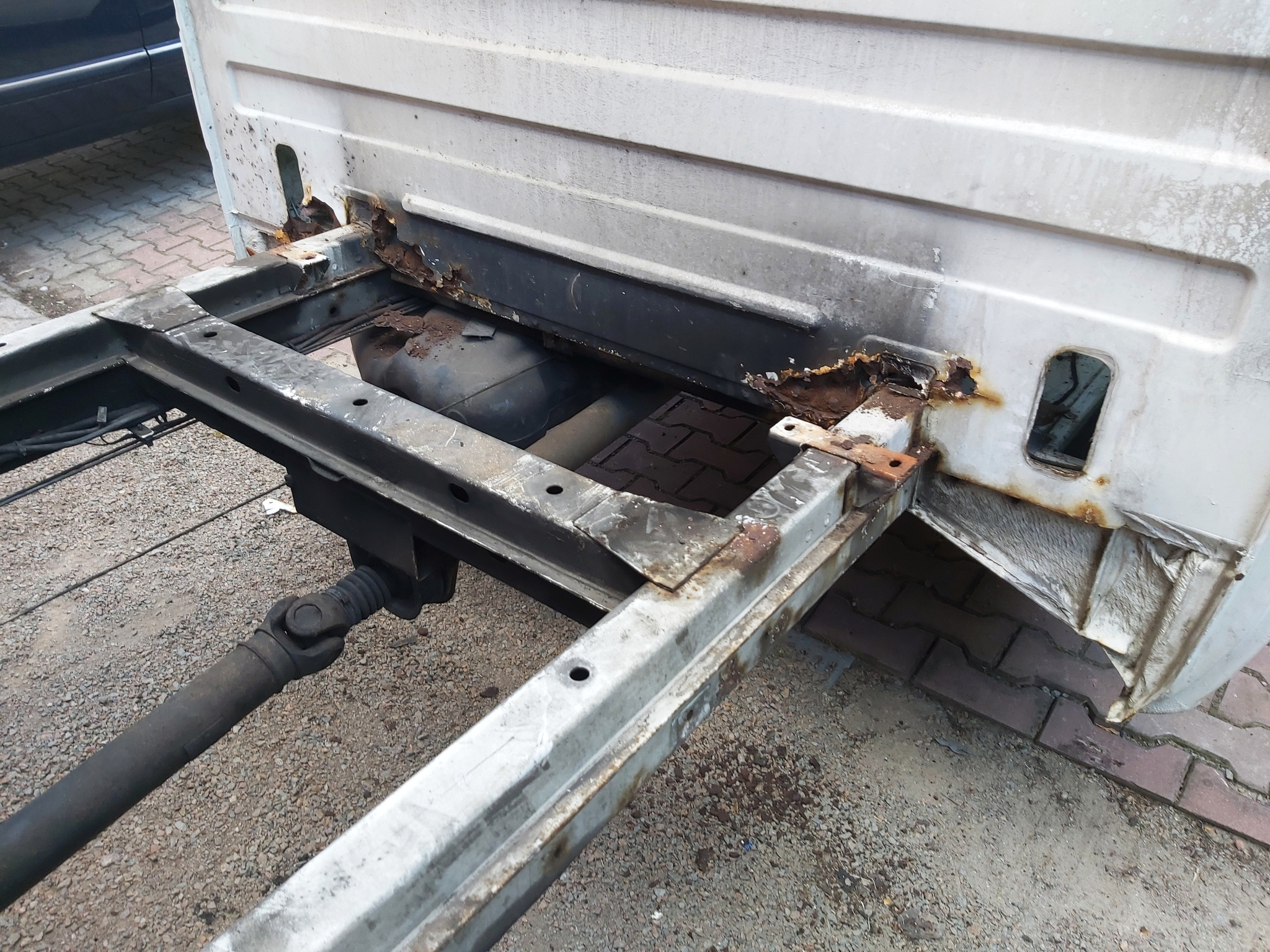 Fragment skorodowanej ramy samochodu dostawczego z widocznym wałem napędowym i elementami karoserii, ukazujący zaawansowany stopień korozji.