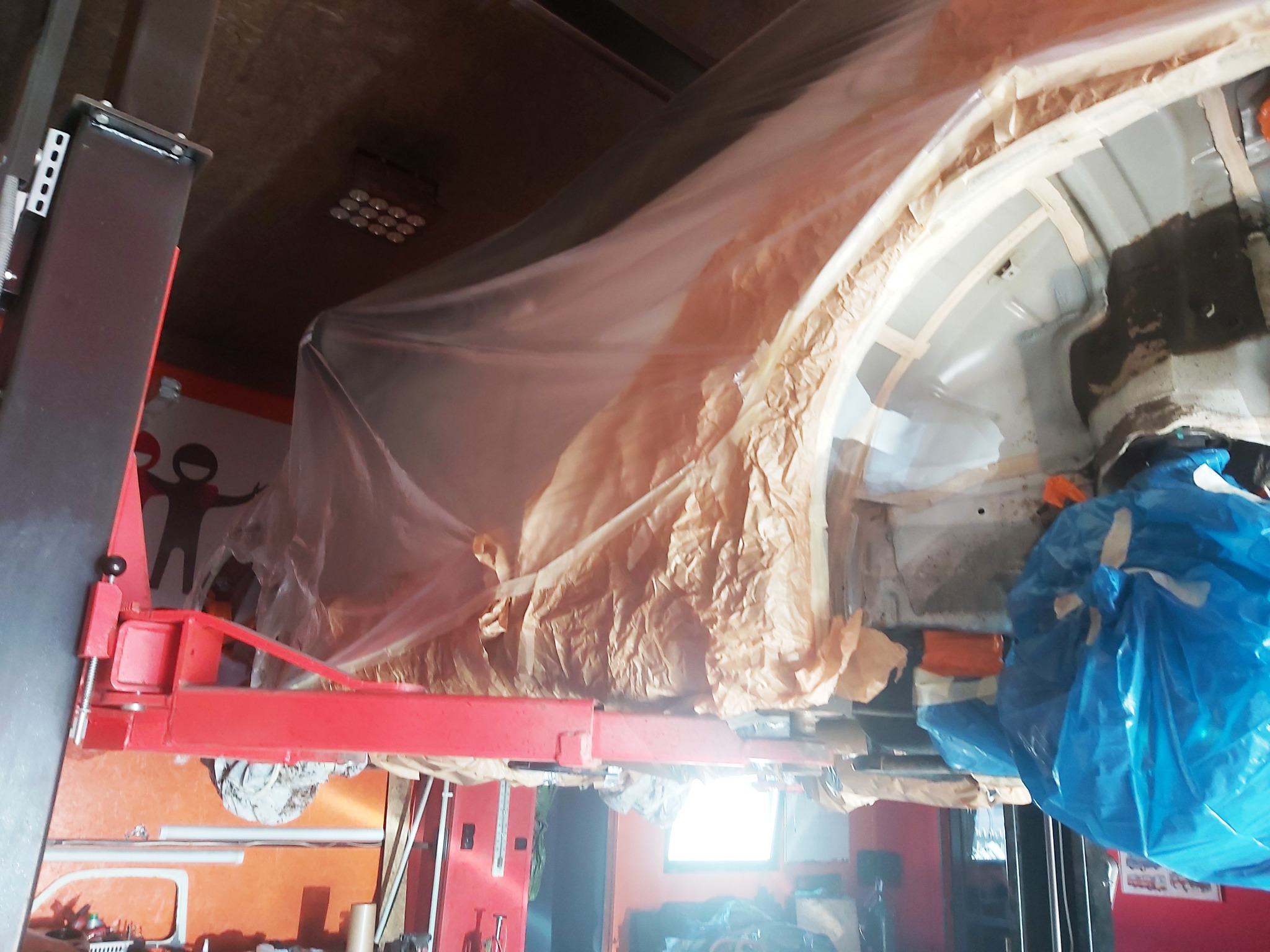 Podwozie samochodu na podnośniku warsztatowym, zabezpieczone folią i papierem maskującym przed pracami blacharsko-lakierniczymi, widoczny fragment karoserii i elementy konstrukcyjne.