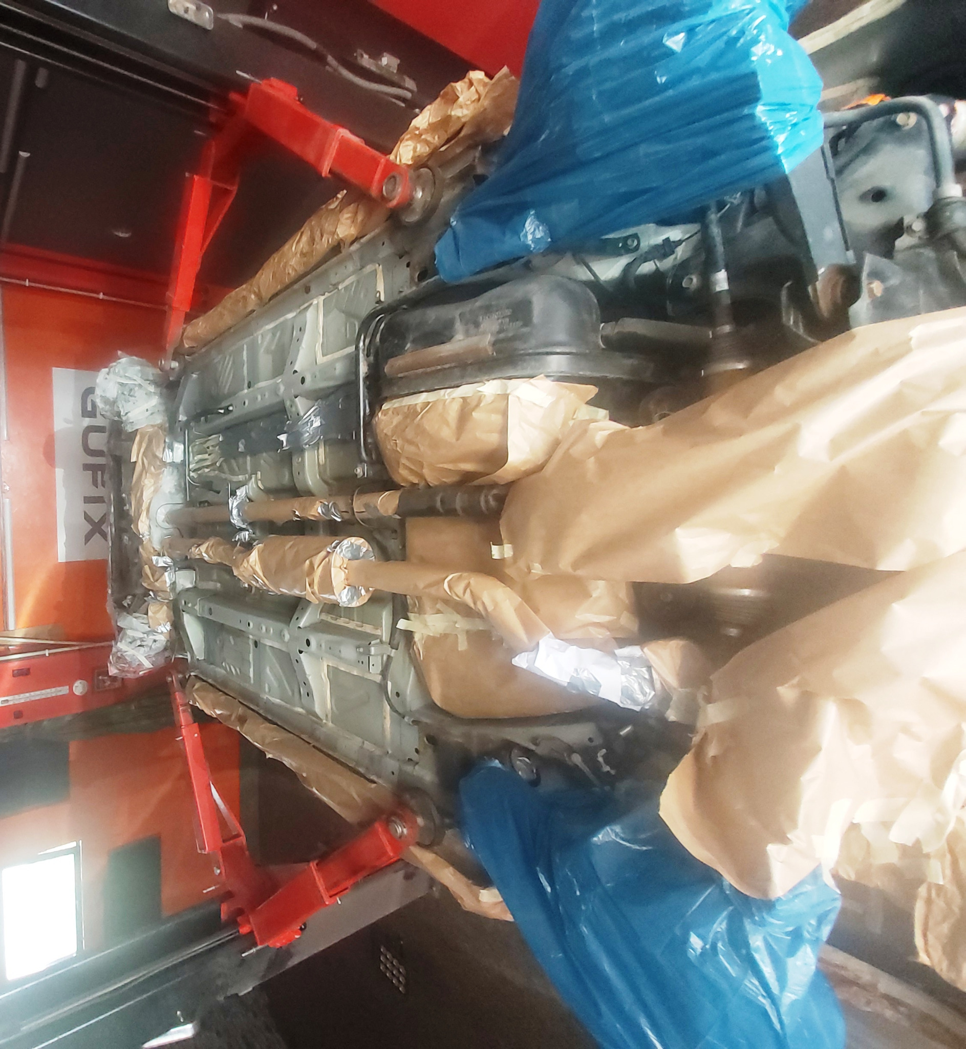 Podwozie samochodu na podnośniku, zabezpieczone papierem i folią przed malowaniem lub konserwacją, widoczne elementy układu wydechowego i podwozia.