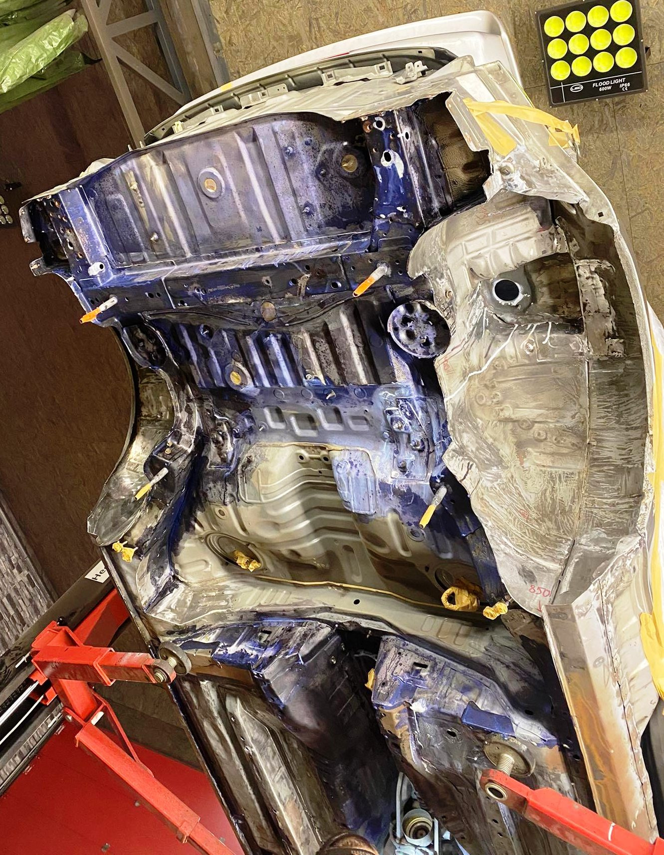 Podwozie samochodu na podnośniku podczas prac blacharskich i lakierniczych, widoczne elementy zabezpieczone taśmą, świeżo nałożona warstwa lakieru w kolorze granatowym.