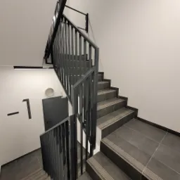 Klatka schodowa z ciemnymi, płytkowanymi schodami z antypoślizgowymi paskami i czarną, metalową balustradą z pionowymi szczeblami. Na ścianie widoczny numer -1 i szare drzwi.