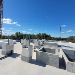 Wznoszenie ścian z bloczków betonowych na dachu budynku w trakcie budowy, widoczny żuraw budowlany i błękitne niebo.