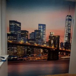 LaDeKo - Fototapeta z widokiem na panoramę Nowego Jorku, w tle niebiesko-pomarańczowe niebo o zmierzchu, na podłodze wiadro z klejem do tapet.