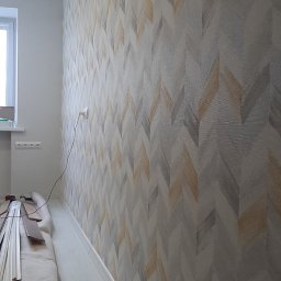 LaDeKo - Ściana z tapetą w jodełkę w odcieniach szarości i beżu, widoczne listwy przypodłogowe i materiały wykończeniowe w rogu pomieszczenia.