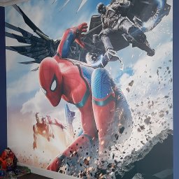 LaDeKo - Fototapeta z motywem superbohaterów Marvela (Spider-Man, Iron Man, Vulture) na ścianie pokoju dziecięcego, widoczna listwa sufitowa, ciemna podłoga, fragment dywanu i poduszki.