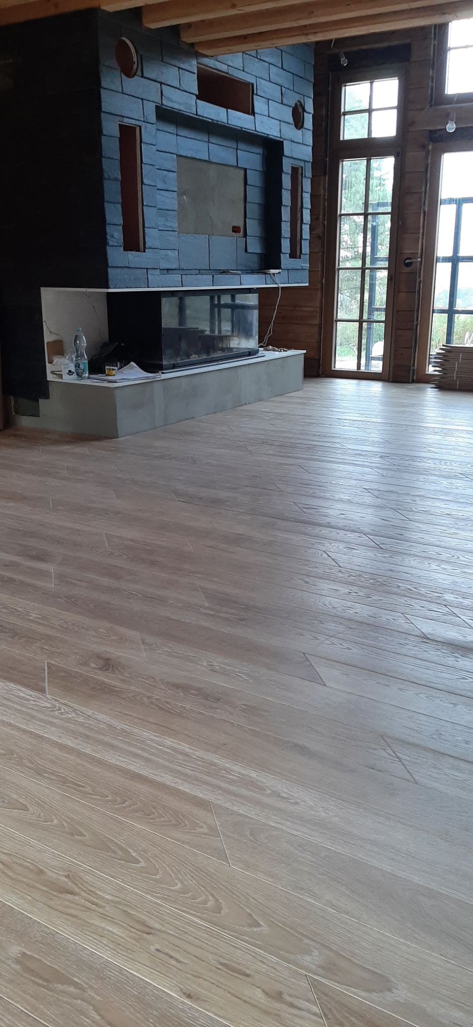 Wnętrze domu w trakcie prac wykończeniowych: jasna drewnopodobna podłoga, kominek obłożony ciemnym kamieniem łupkowym, duże okna z drewnianą stolarką.
