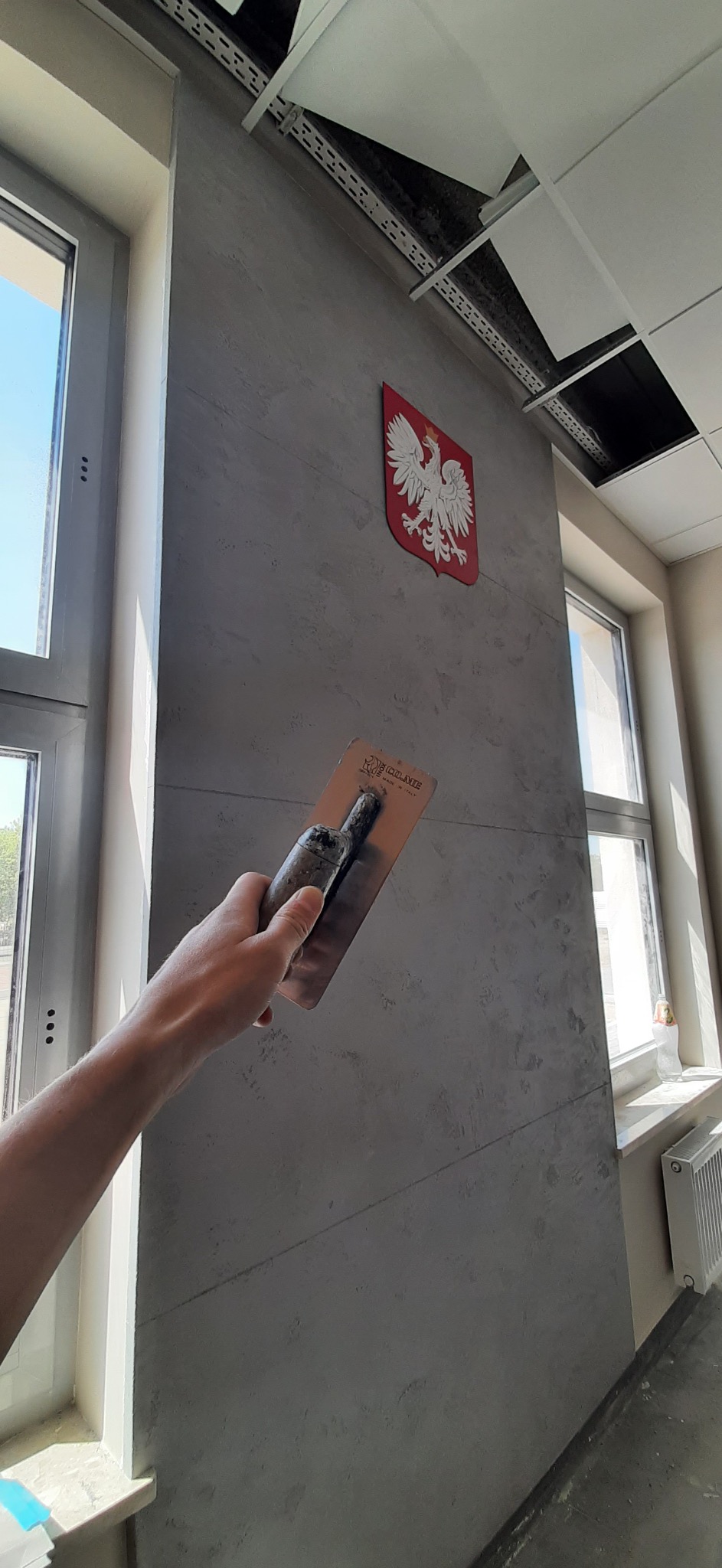 Nakładanie dekoracyjnego tynku w odcieniach szarości na ścianę w pomieszczeniu biurowym, widoczny herb Polski i fragment okna.