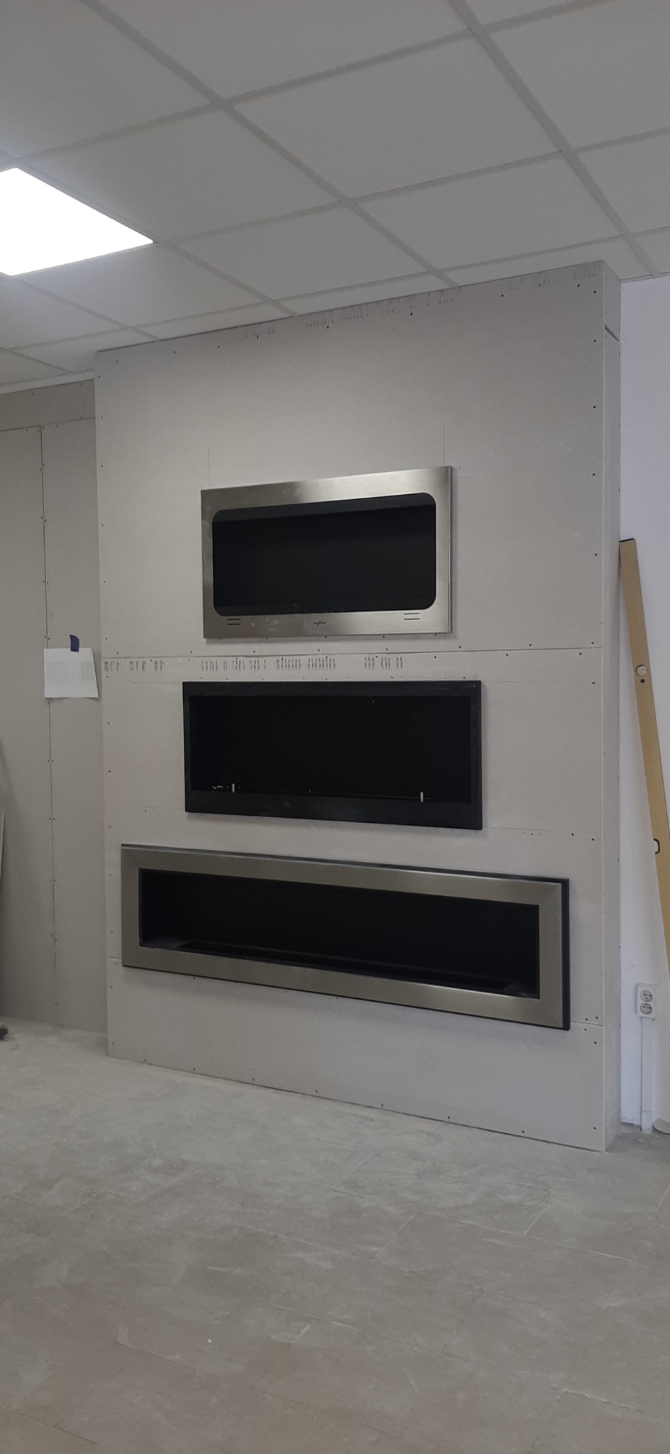 Ściana z płyt kartonowo-gipsowych z zamontowanymi trzema wkładami kominkowymi o różnych rozmiarach, w trakcie montażu, widoczny poziomica oparta o ścianę.