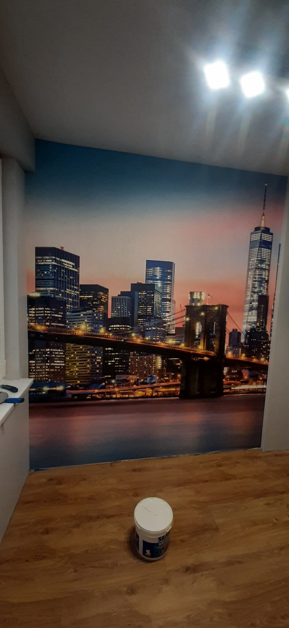 Fototapeta z widokiem na panoramę Nowego Jorku, w tle niebiesko-pomarańczowe niebo o zmierzchu, na podłodze wiadro z klejem do tapet.