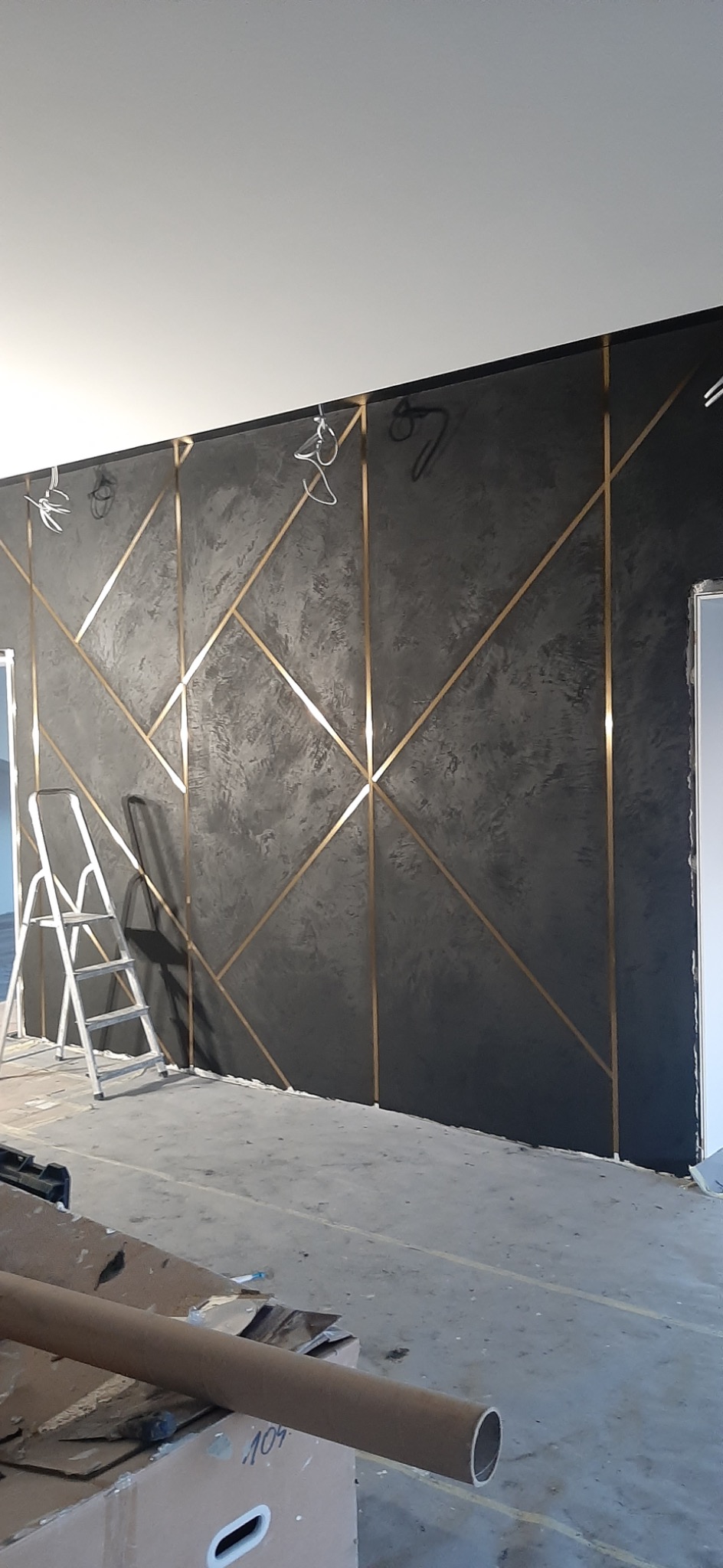 Ściana pokryta szarą, fakturowaną farbą z geometrycznym wzorem wykonanym ze złotych listew, widoczna aluminiowa drabina i elementy instalacji elektrycznej u góry.