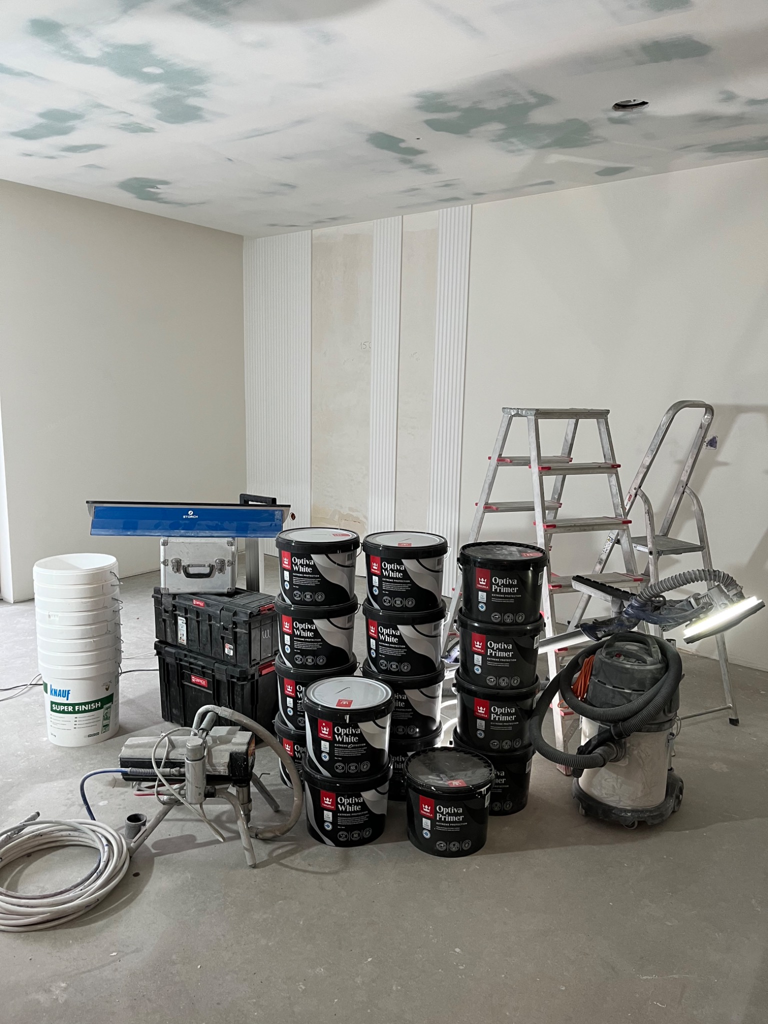 Przygotowania do malowania wnętrza: wiadro z gładzią, agregat malarski, puszki z farbą Optiva White i Primer, szlifierka do gipsu z odkurzaczem, drabiny, narzędzia, sufit z widocznymi łatami...