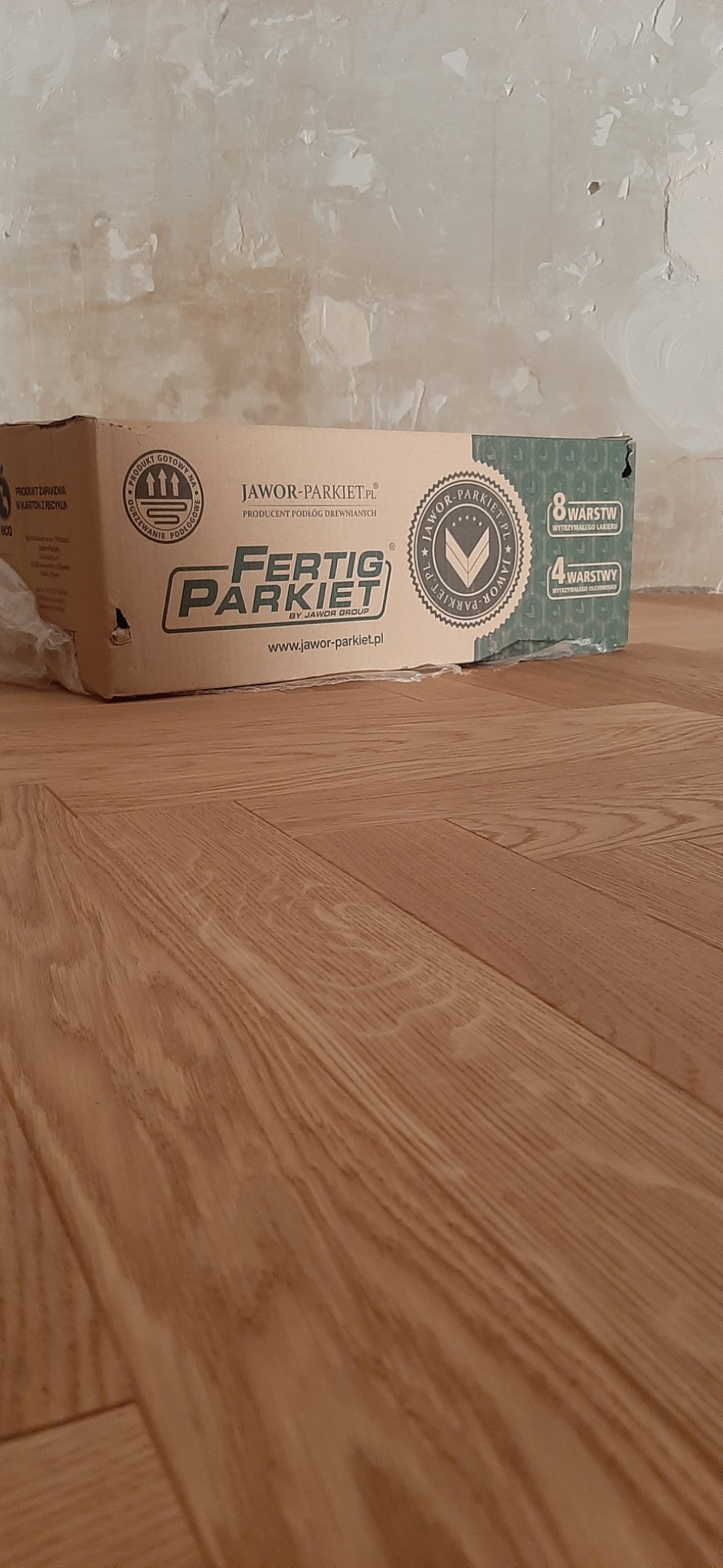 Ułożony parkiet drewniany w jodełkę, w tle karton z napisem 'Fertig Parkiet' i surowa ściana przed wykończeniem.