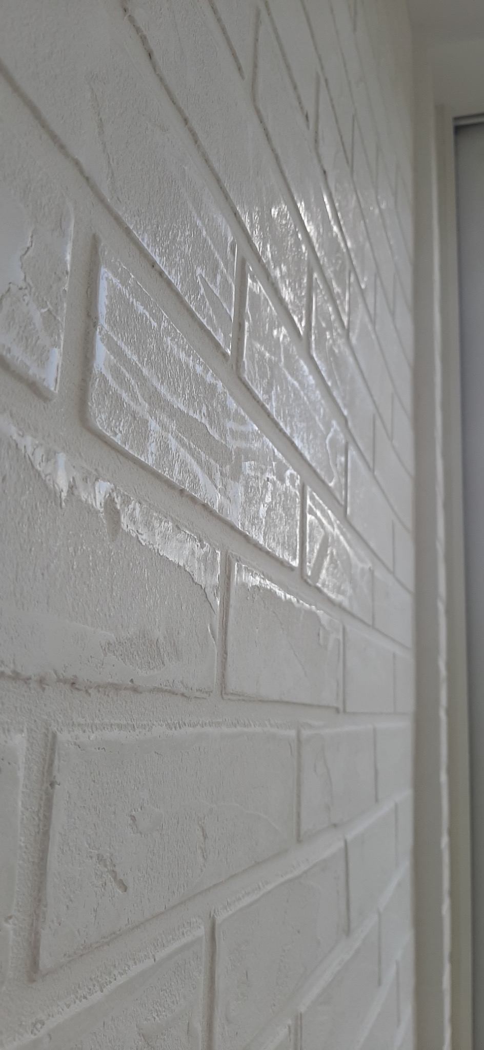 Ściana wykończona białą, strukturalną farbą imitującą cegłę, widoczna z boku, z delikatnym połyskiem i widocznymi nierównościami faktury.