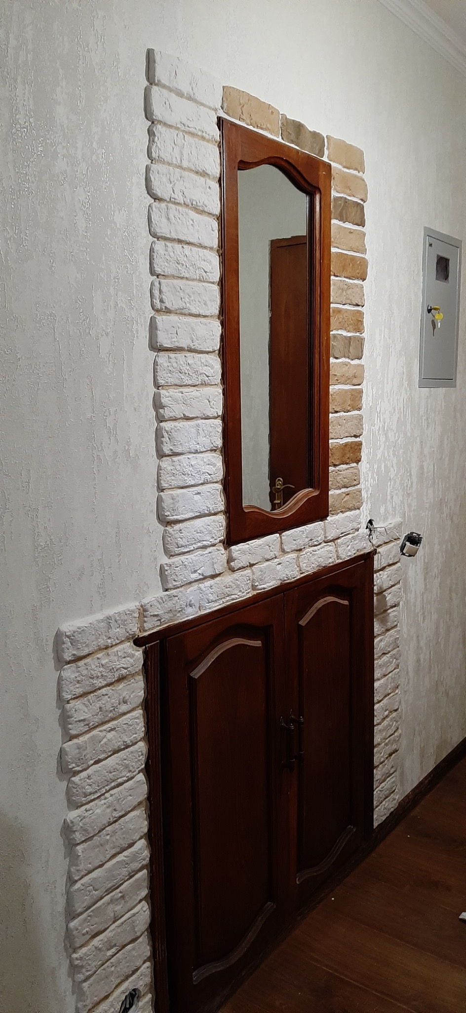 Narożnik ściany wykończony dekoracyjną cegłą w odcieniach bieli i beżu, obudowa z ciemnego drewna z lustrem w ramie, widoczny fragment instalacji elektrycznej.