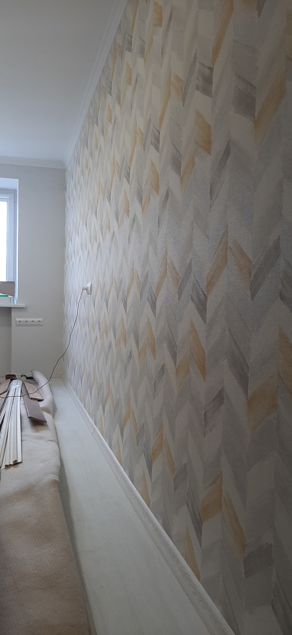 Ściana z tapetą w jodełkę w odcieniach szarości i beżu, widoczne listwy przypodłogowe i materiały wykończeniowe w rogu pomieszczenia.