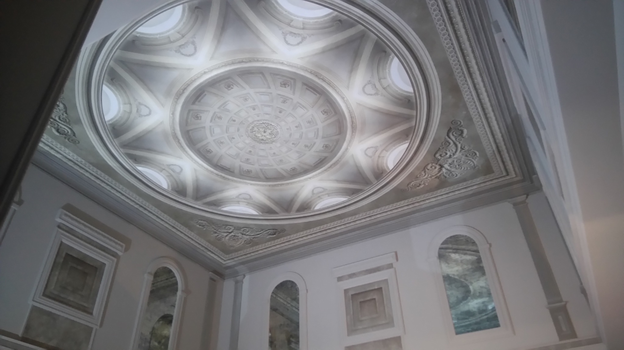 Kopuła sufitowa z dekoracyjnym wzorem w odcieniach szarości, widoczna z dołu, z elementami sztukaterii i okrągłymi oknami w górnej części.