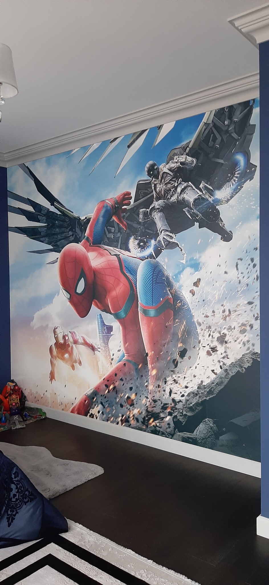 Fototapeta z motywem superbohaterów Marvela (Spider-Man, Iron Man, Vulture) na ścianie pokoju dziecięcego, widoczna listwa sufitowa, ciemna podłoga, fragment dywanu i poduszki.