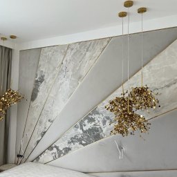 Bild in Sp.zo.o. - Wykończona ściana za łóżkiem w sypialni z dekoracyjnymi panelami w odcieniach szarości i bieli, imitującymi marmur, z złotymi akcentami oraz wiszące złote lampy.