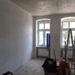 Pokój w trakcie prac wykończeniowych: świeżo pomalowane ściany w odcieniach szarości, dwa okna z niebieskimi zasłonami, drabiny malarskie, wiadro z odpadami budowlanymi i prowizoryczny stolik...