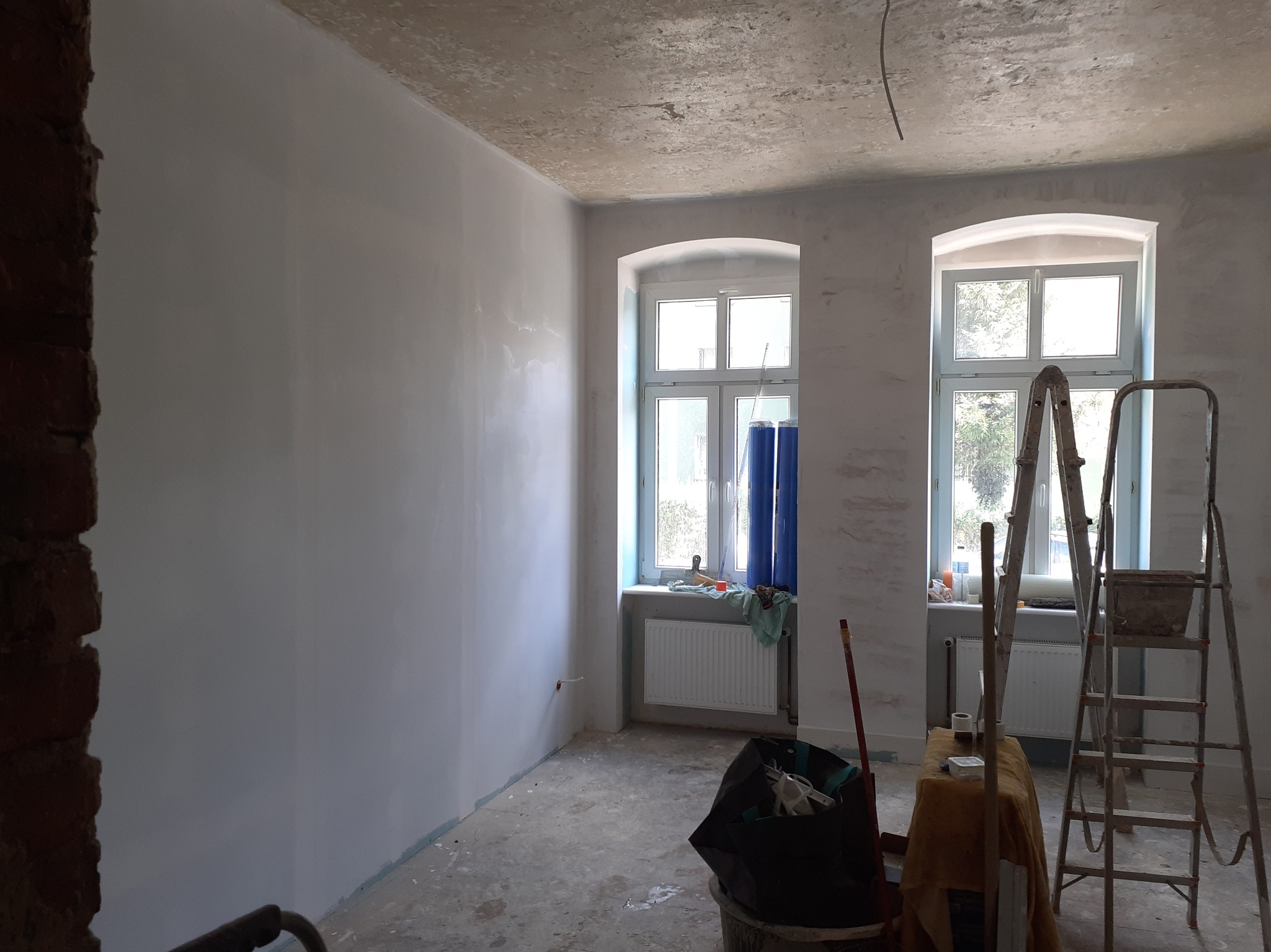 Pokój w trakcie prac wykończeniowych: świeżo pomalowane ściany w odcieniach szarości, dwa okna z niebieskimi zasłonami, drabiny malarskie, wiadro z odpadami budowlanymi i prowizoryczny stolik...
