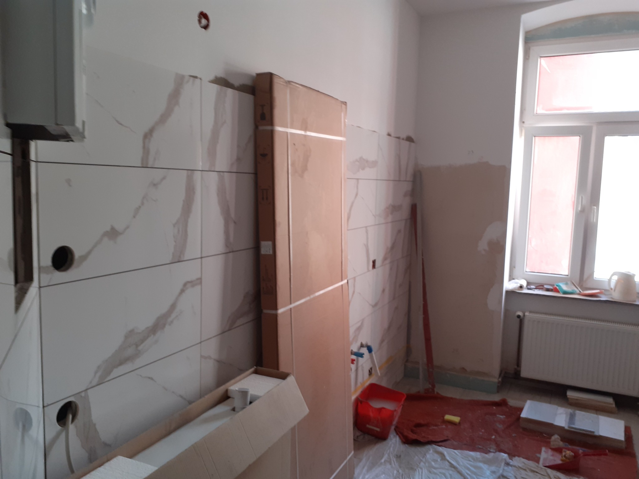 Wnętrze w trakcie remontu: ułożone płytki imitujące marmur na ścianie, zapakowane drzwi oparte o ścianę, czerwone wiadro z wodą, zabezpieczona folią podłoga, grzejnik pod oknem.