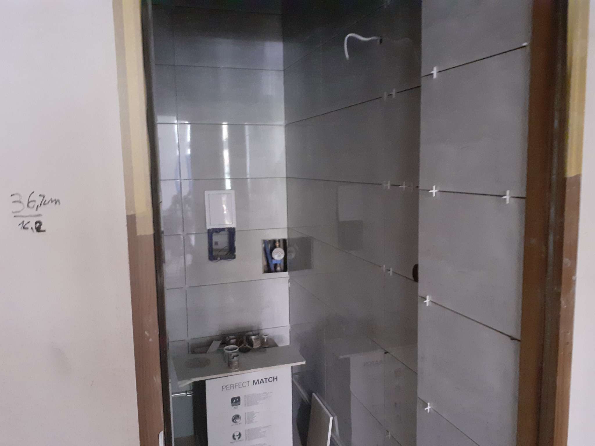 Łazienka w trakcie wykańczania: szare płytki ułożone do pewnej wysokości, widoczne krzyżyki dystansowe, przygotowane otwory na armaturę, częściowo zasłonięte opakowanie płytek z napisem 'Perfect...