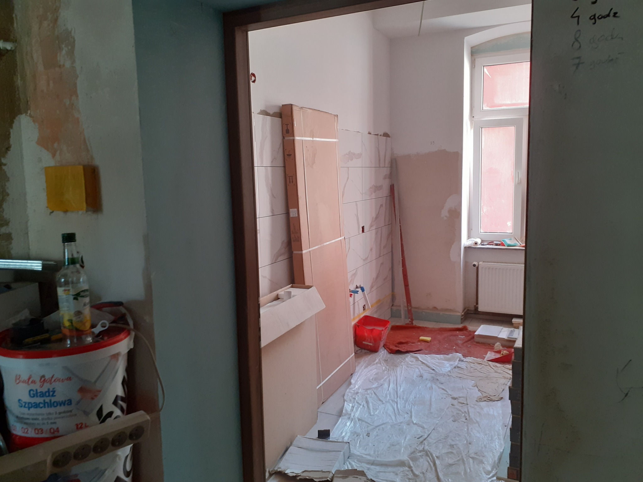 Wnętrze w trakcie remontu: częściowo położone płytki, zabezpieczona podłoga folią, wiadro z gładzią szpachlową, okno z grzejnikiem. Widoczne ślady po gładzi na ścianach.