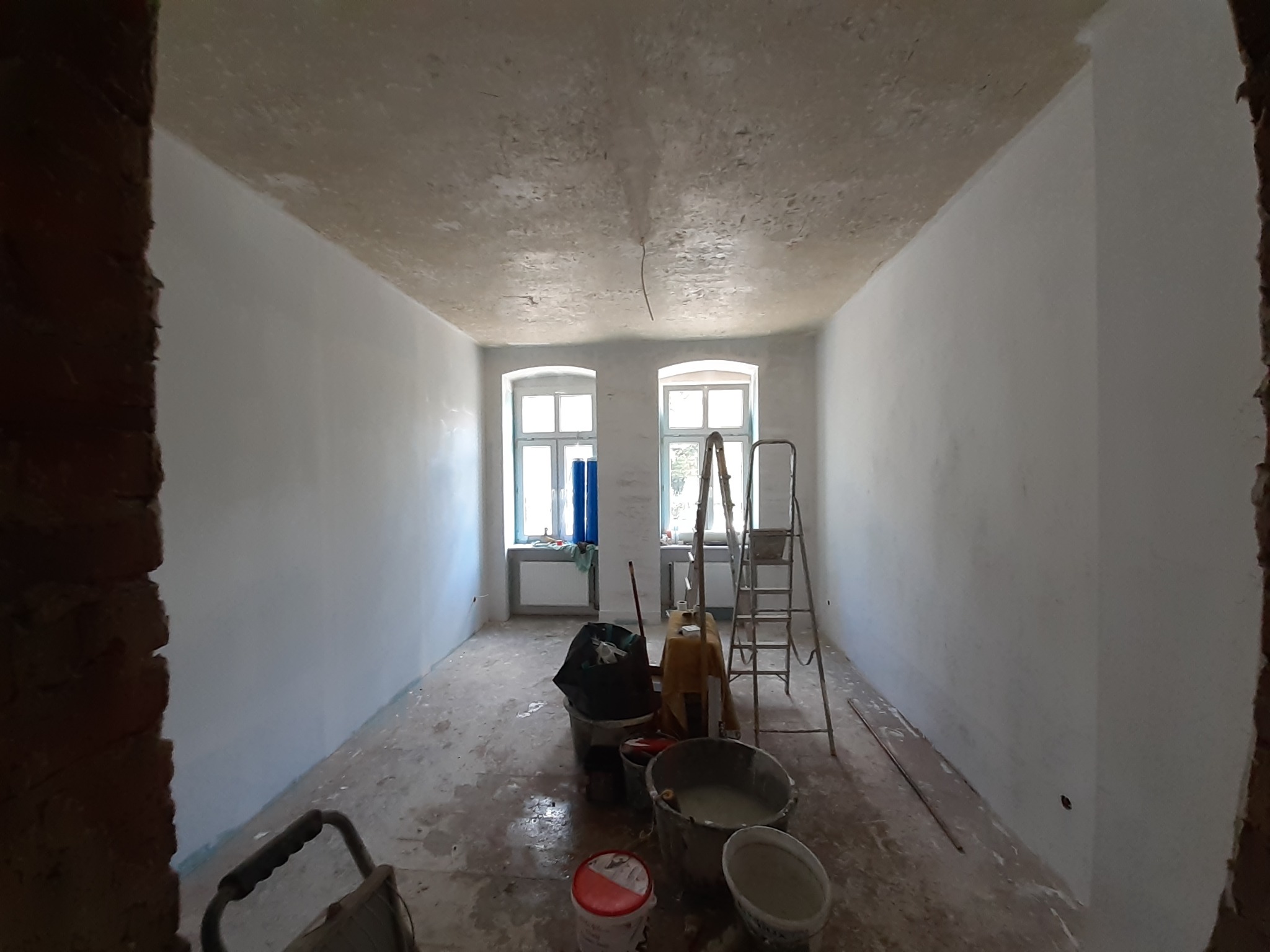 Pokój w trakcie odświeżania, ściany częściowo pomalowane na biało, widoczne drabiny, wiadra z farbą i narzędzia malarskie, sufit z widocznymi śladami starej farby.