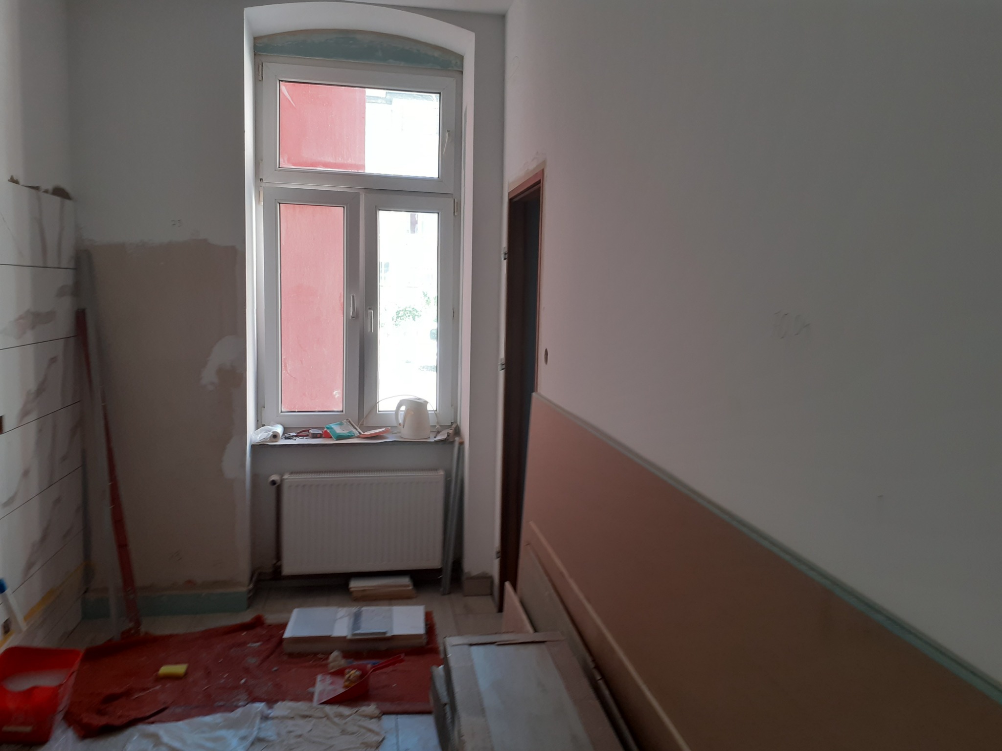 Pokój w trakcie prac wykończeniowych: częściowo położone płytki imitujące marmur, przygotowane panele ścienne, okno z grzejnikiem, ślady po szpachlowaniu na ścianie.
