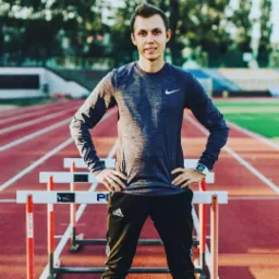 Młody mężczyzna w sportowym stroju Nike i Adidas, stojący na bieżni lekkoatletycznej z przeszkodami, z zegarkiem sportowym na nadgarstku.
