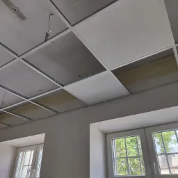 Montaż sufitu podwieszanego kasetonowego z widocznymi elementami konstrukcyjnymi i częściowo zamontowanymi panelami, wnętrze z oknami w tle.