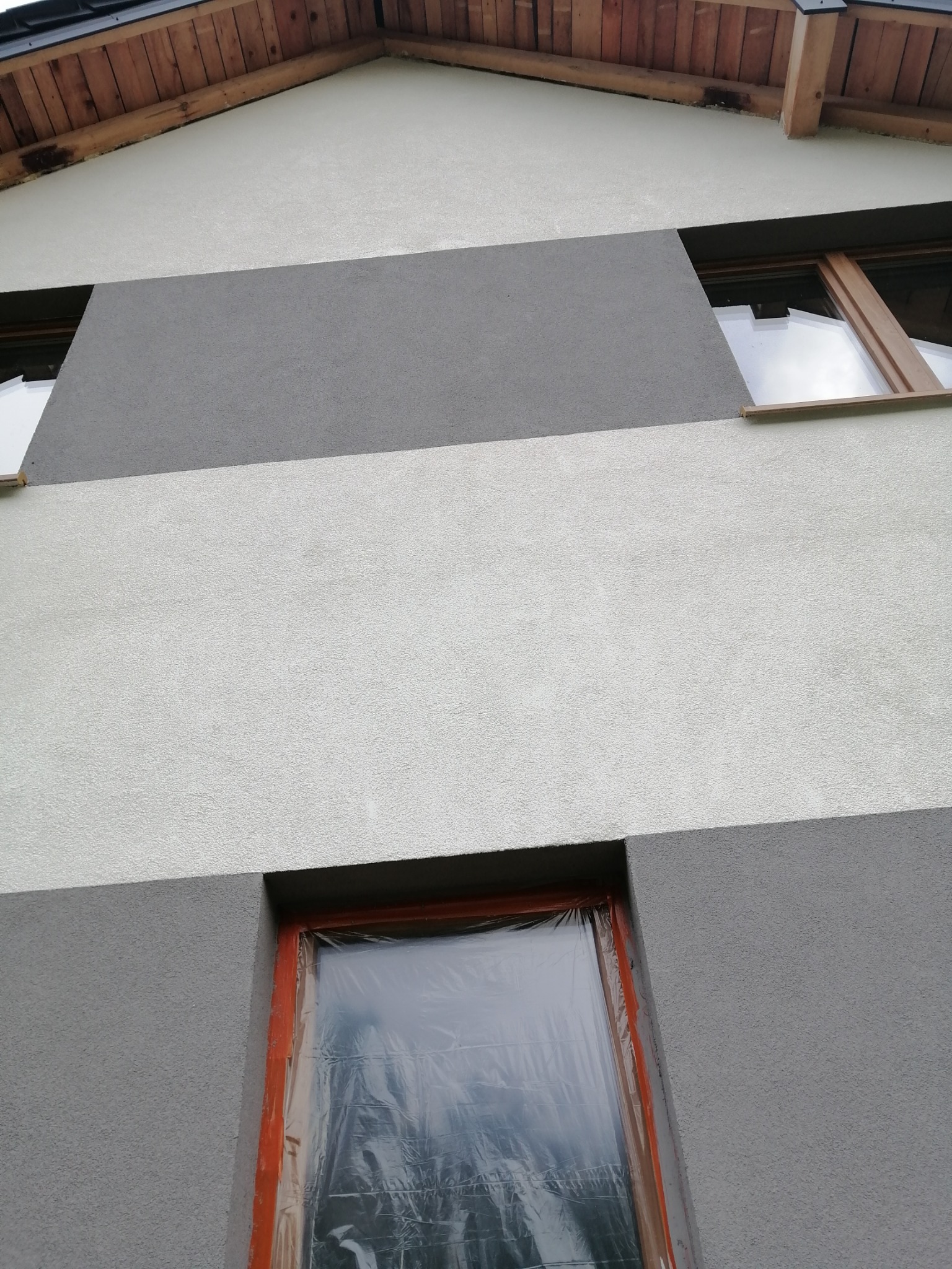 Fragment elewacji budynku z poziomymi pasami w odcieniach szarości, okna zabezpieczone folią malarską, widoczny fragment drewnianej konstrukcji dachu.