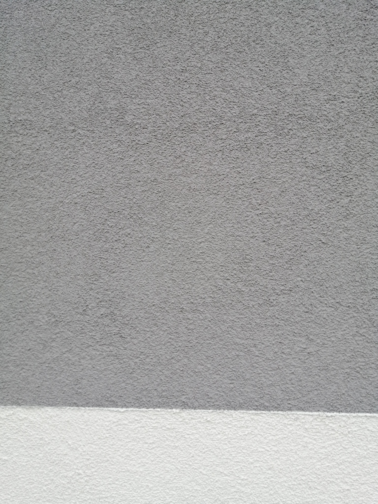 Szara elewacja z wyraźną, ziarnistą strukturą tynku, oddzielona poziomą białą linią od dolnej części ściany.