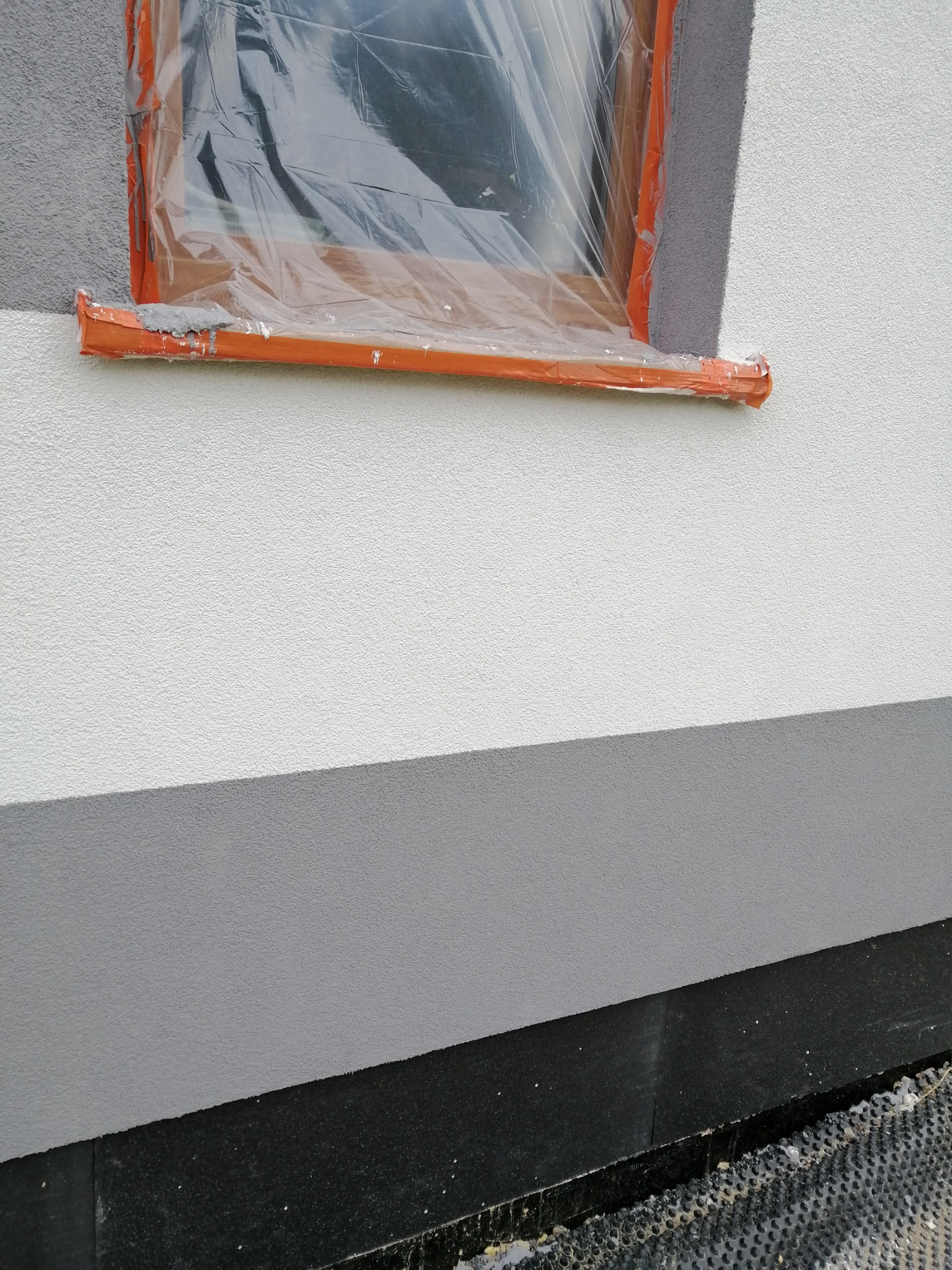 Elewacja budynku z oknem zabezpieczonym folią i taśmą malarską, widoczny fragment pomalowanej ściany na biało i szaro, dolna część elewacji wykończona czarnym materiałem.