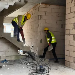 Dwóch pracowników w kaskach i kamizelkach odblaskowych, jeden używa młota pneumatycznego na betonowych schodach, drugi przenosi gruz w surowym wnętrzu budynku z białych bloków.