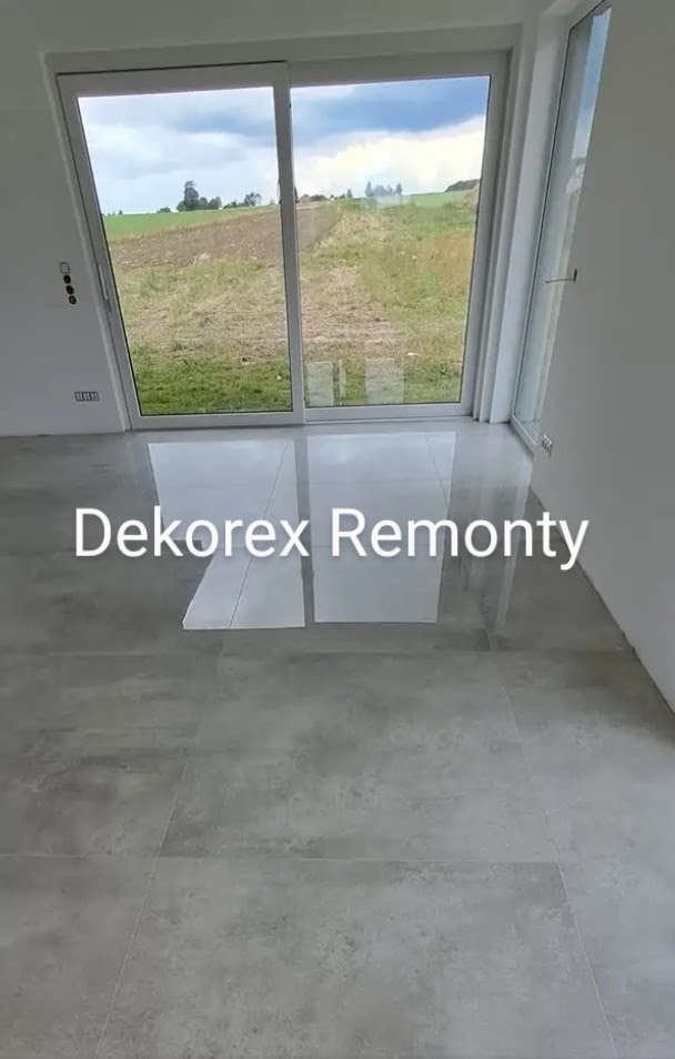 Wykończona podłoga z szarych płytek w jasnym pomieszczeniu z dużym oknem przesuwnym z widokiem na zielone pole. Widoczne odbicie światła na płytkach.