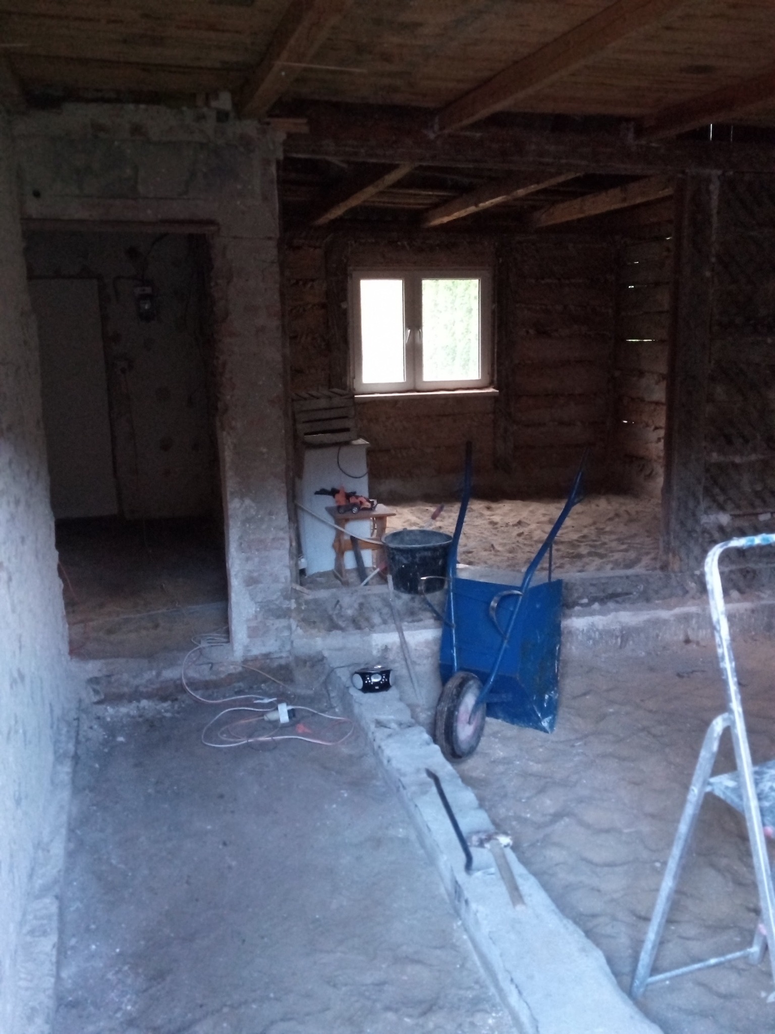 Wnętrze w trakcie remontu: widoczne surowe ściany, drewniany strop, okno, taczka, drabina oraz narzędzia budowlane, przewody elektryczne.