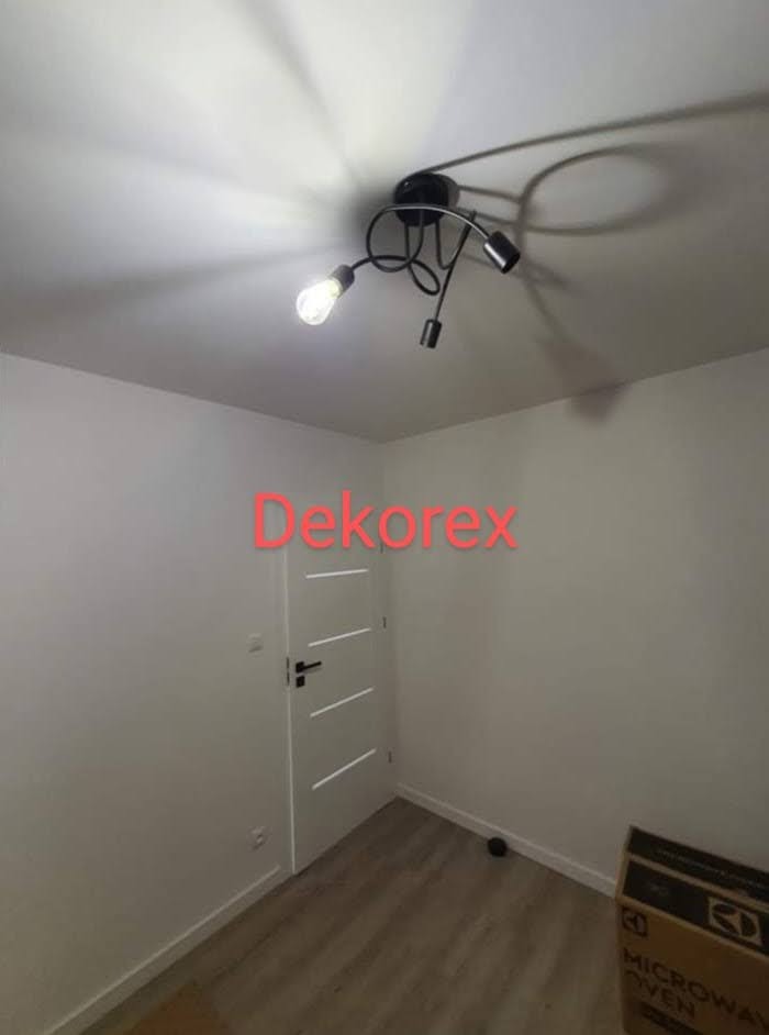 Świeżo pomalowane na biało wnętrze pokoju z nowoczesną lampą sufitową z trzema żarówkami, widoczne drzwi wewnętrzne i kartonowe pudło na podłodze.