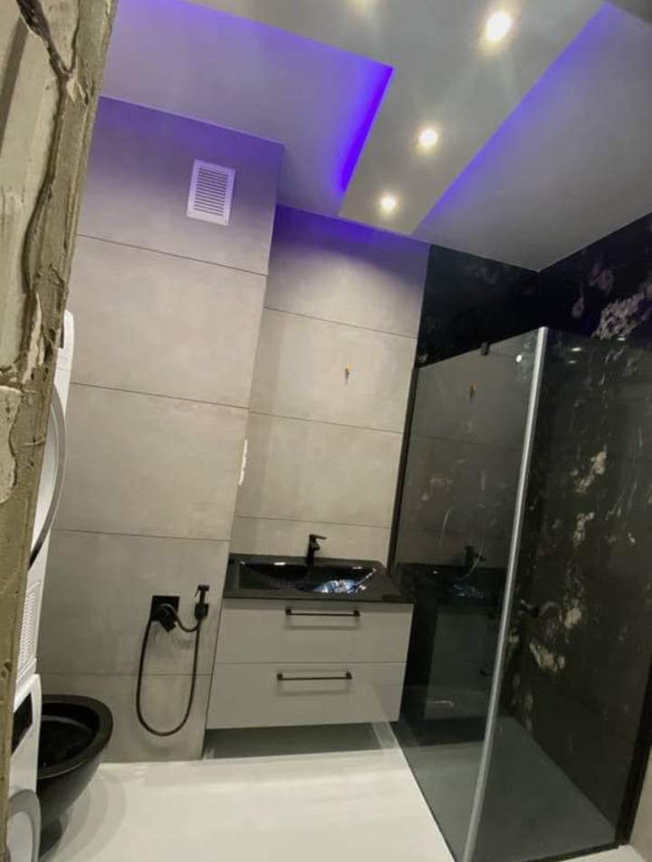 Nowoczesna łazienka z szarą płytką ścienną, czarną armaturą, podświetlanym sufitem LED w kolorze fioletowym i kabiną prysznicową z czarnego marmuru.
