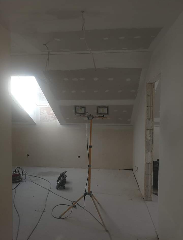 Wnętrze pokoju ze skośnym oknem dachowym w trakcie montażu płyt kartonowo-gipsowych, widoczne kable, narzędzia i lampa na statywie.