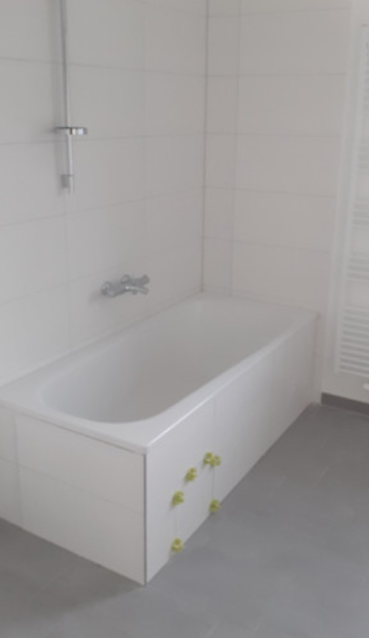 Biała wanna w obudowie z płytek, świeżo po montażu, z zielonymi krzyżykami dystansowymi widocznymi na dolnej krawędzi, szare płytki podłogowe i białe płytki ścienne, widoczny grzejnik drabinkowy.