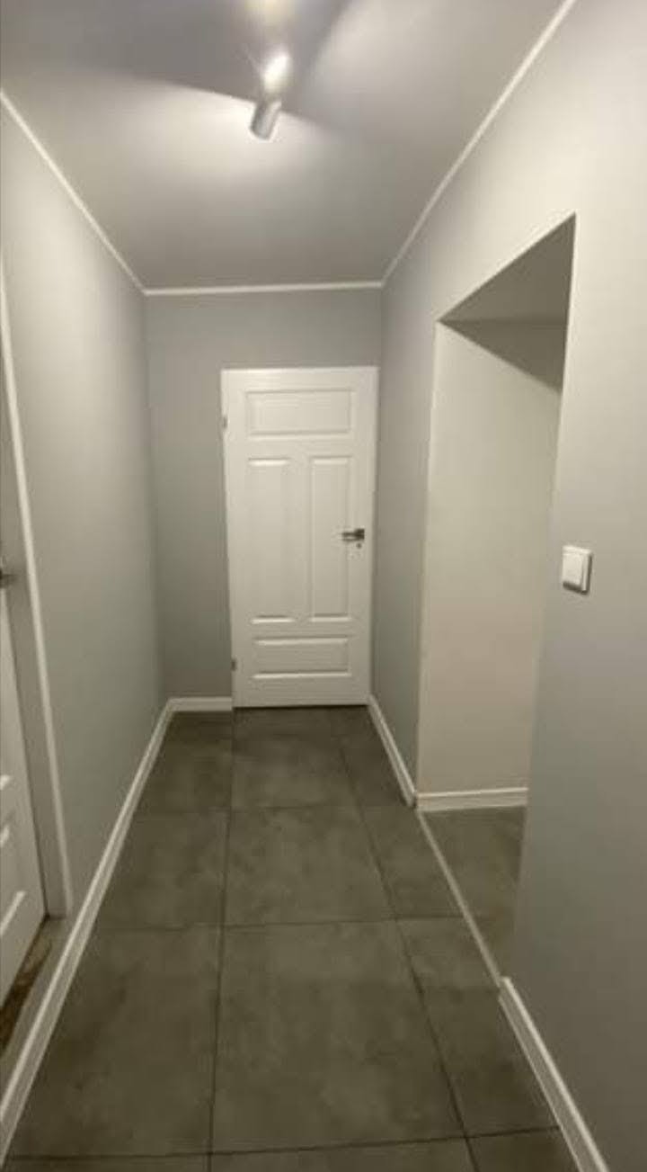 Wnętrze korytarza z szarymi ścianami, ciemnymi płytkami podłogowymi i białymi drzwiami, oświetlone pojedynczym reflektorem sufitowym.