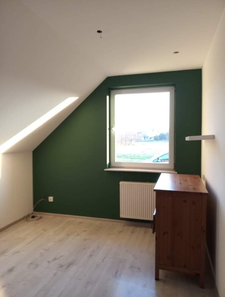 Pokój na poddaszu po remoncie: ściana pomalowana na ciemnozielono, jasna podłoga, drewniana komoda przy ścianie z grzejnikiem pod oknem.