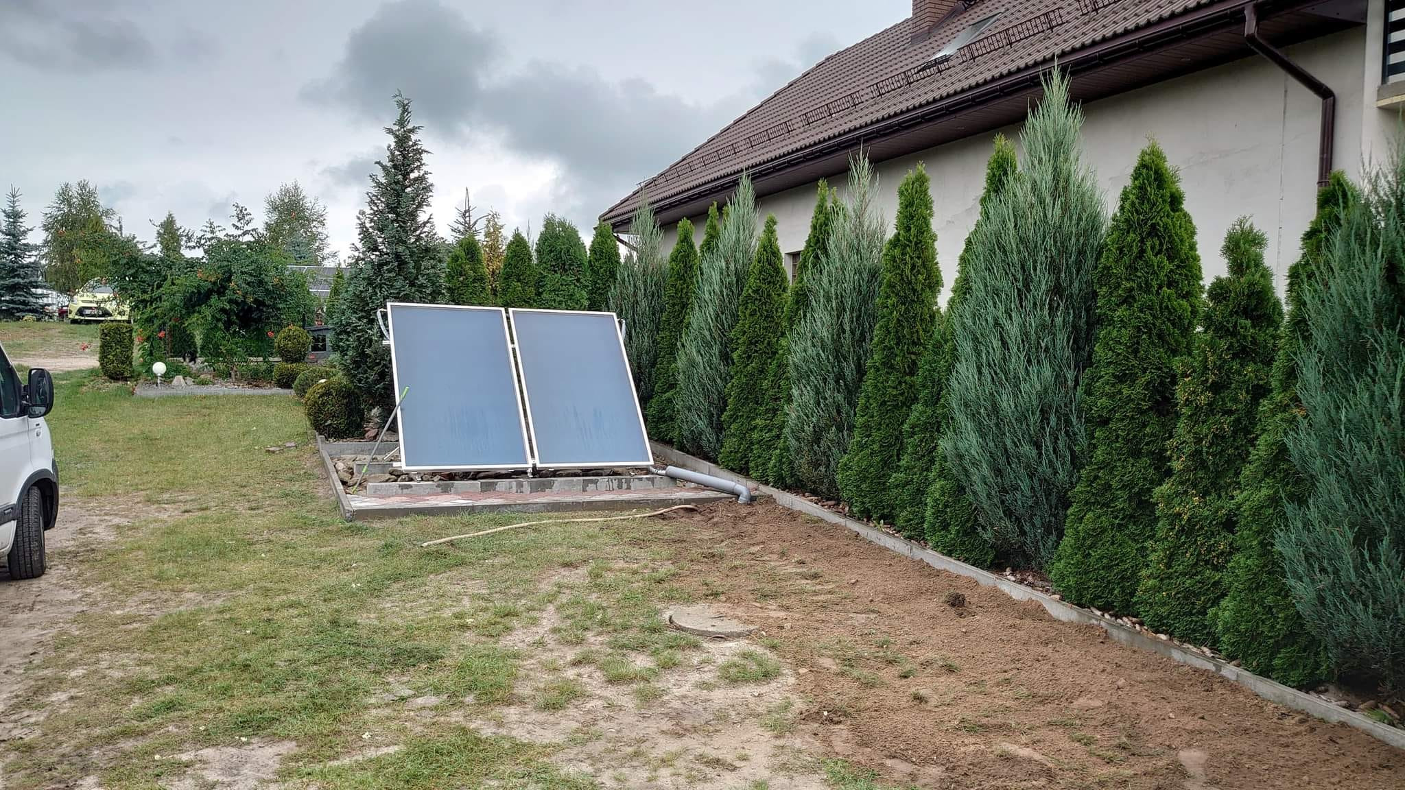 Dwa panele słoneczne zamontowane na niskim betonowym fundamencie, z widocznym szarym przewodem odprowadzającym wodę, na tle rzędu wysokich, zielonych tuj i ściany budynku z brązowym dachem.