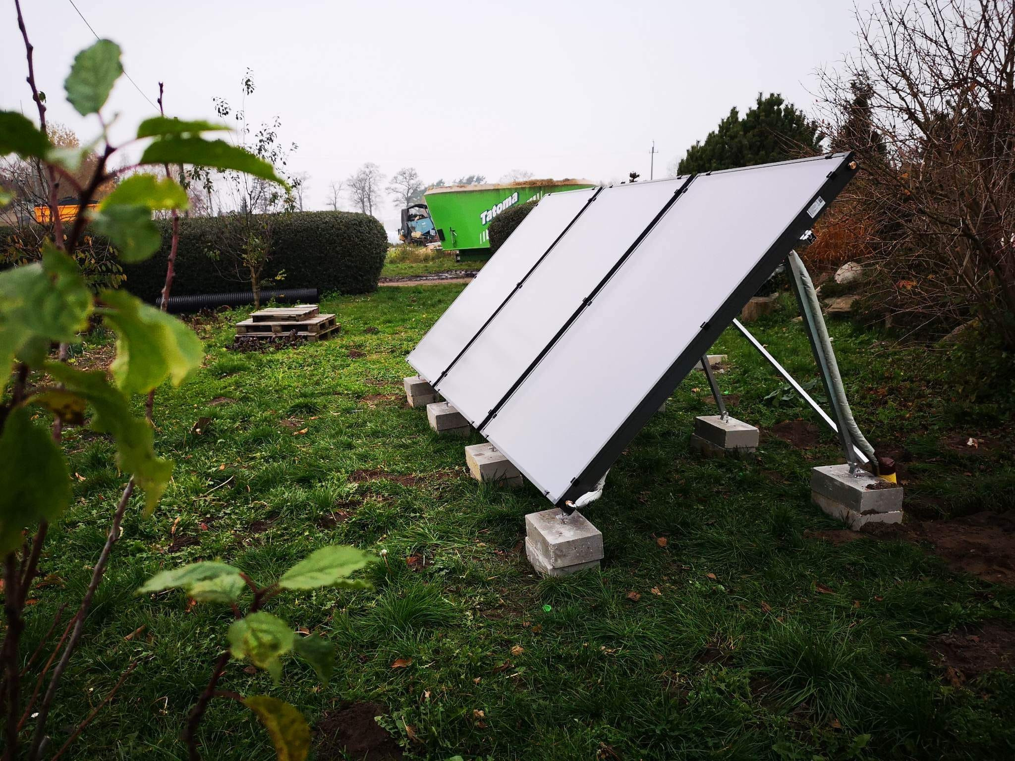 Trzy panele słoneczne ustawione na trawniku, wsparte na betonowych blokach, w tle zielona przyczepa rolnicza z napisem 'Tatoma'.